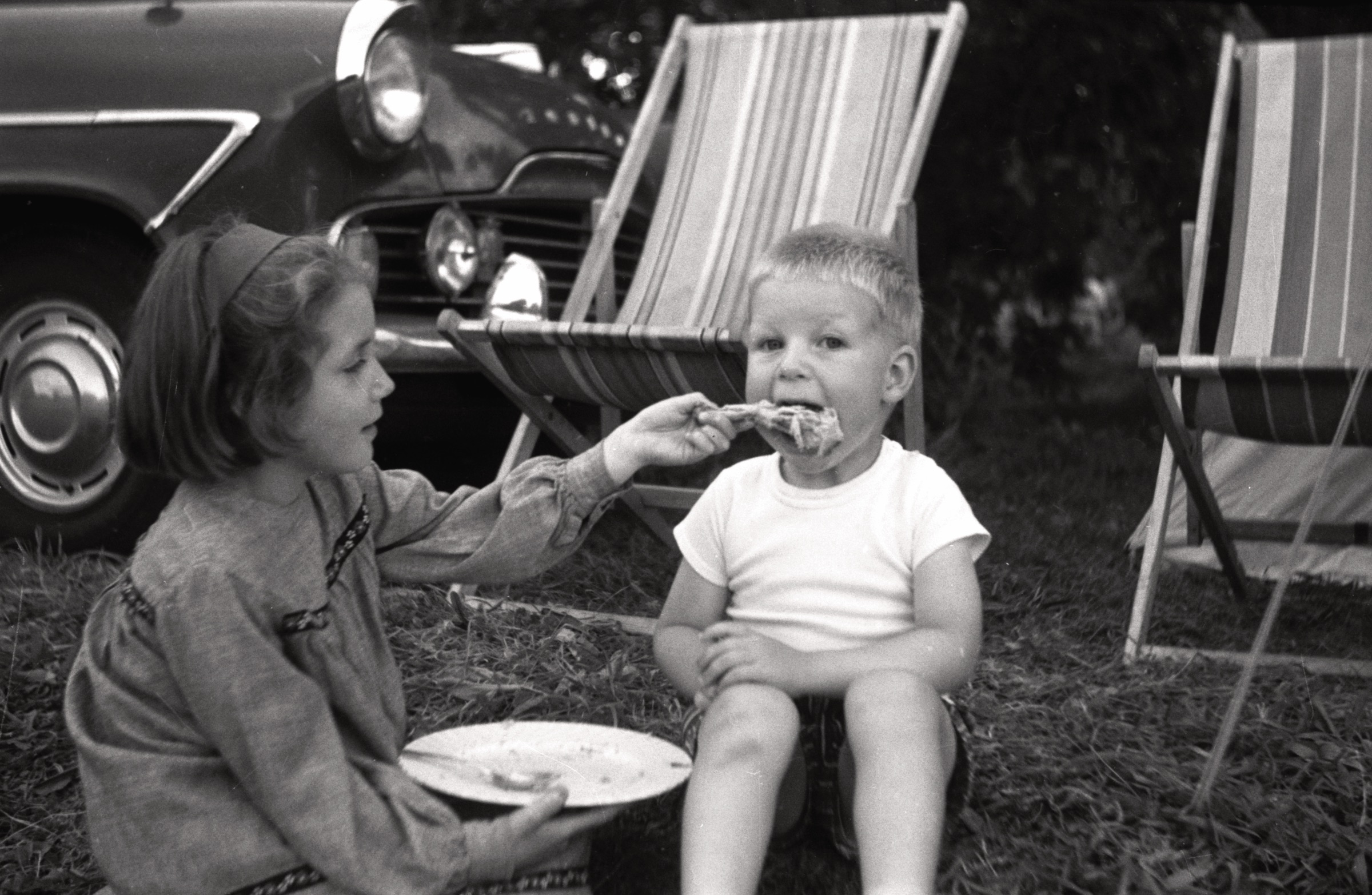 Brigita Lieģis baro Rolandu Blezūru, LNPL Latviešu māja Almelijā, Herefordšīrā, 1970. gadu vidus. Fonā Jāņa Veisa automašīna 'Ford Zodiac'.