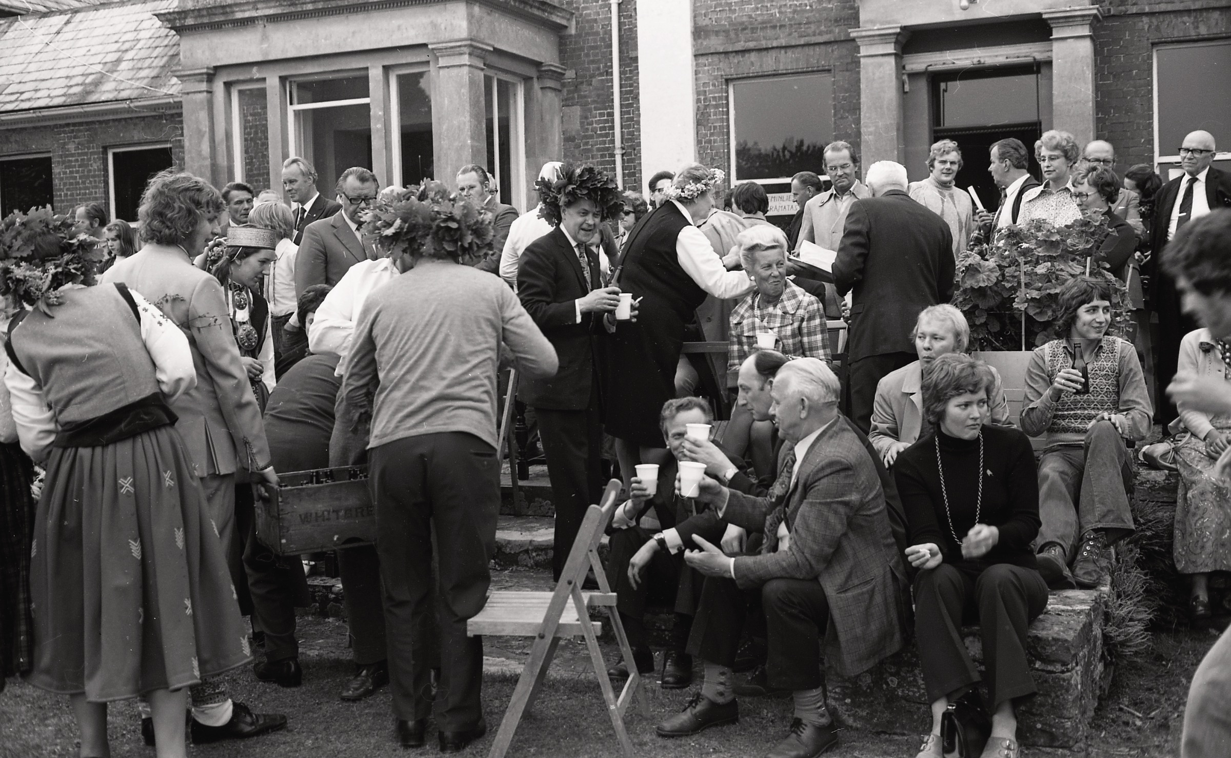 Jāņu svinēšana LNPL Latviešu mājā Almelijā, Herefordšīrā, 1973. gada 30. jūnijā. Aizmugurē no labās ar brillēm Austra Freimane, Maija Bisenieks, Valdis Priede sarunā ar Bruno Deksni. Vidū ar vainagu Leopolds Rumba un Marija Ķeņge (ar vainagu). Viņiem pa vidu ar tumšām brillēm Erna Balode. No labās pie loga Valdis Liepiņš, Imants Pālītis un Jānis Tauriņš. Priekšā no labās ar pudeli Imants Lieģis un Imants Lūsis, starp abiem Ruta Lūsis. Priekšā no kr. tautas tērpā Jānis Prauliņš.