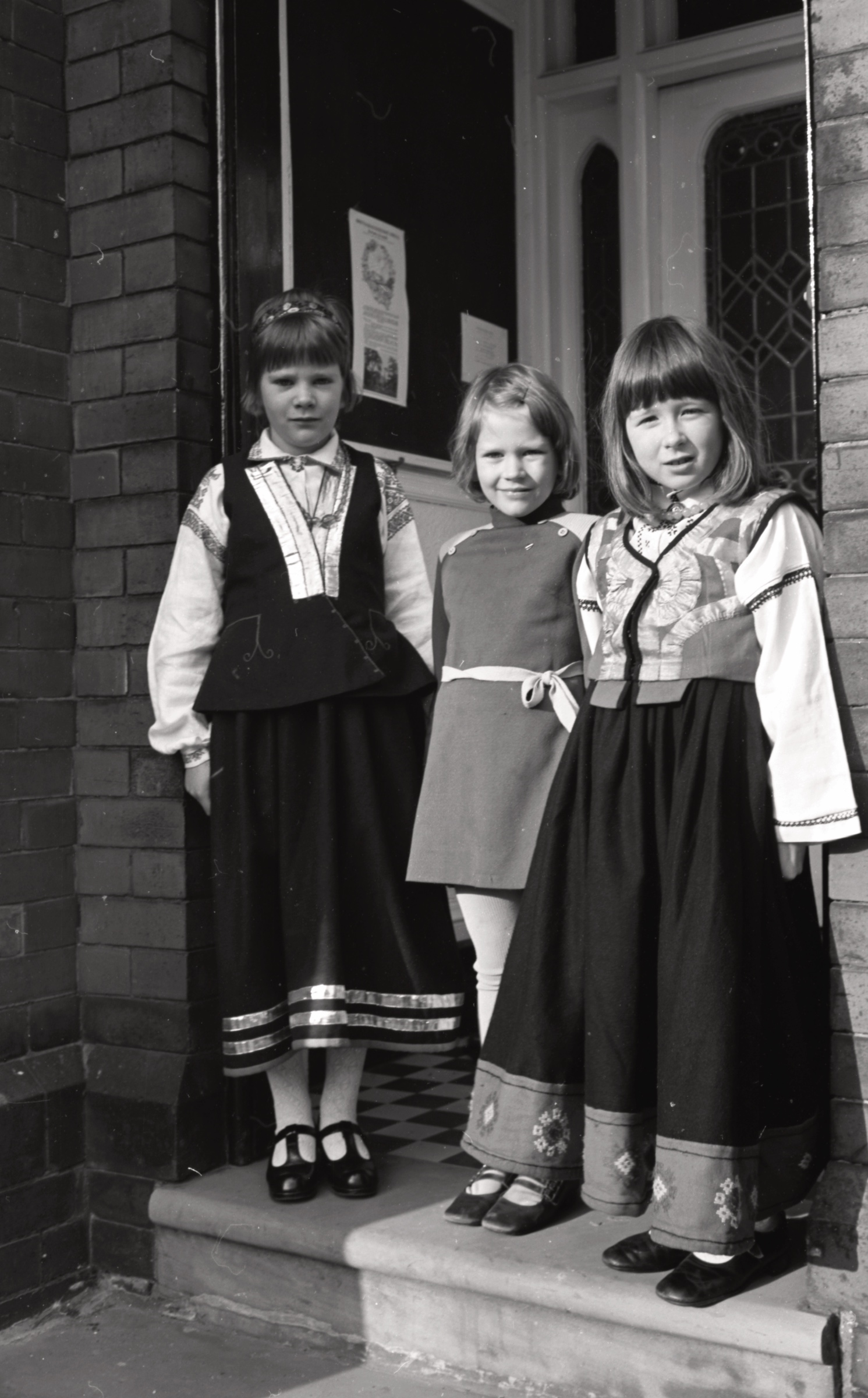 Māsas Dace, Zane un Anita Zaķis tautas tērpos pie DVF Boltonas nodaļas jauniegādā nama ieejas durvīm, Boltona, ap 1974. gadu.