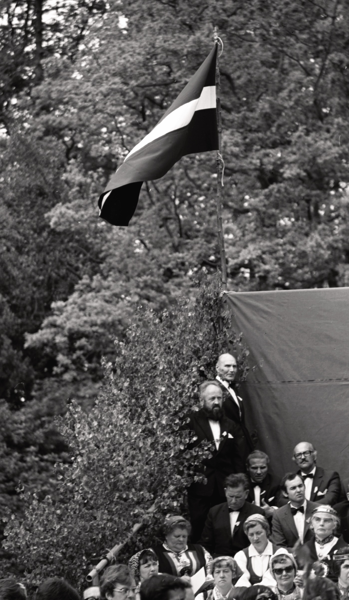 Zem Latvijas karoga. Kopkoris 1. Anglijas latviešu brīvdabas dziesmu dienās 1974. gada 26. maijā Londonas latviešu luterāņu draudzes īpašumā Rofantas muižā, Saseksas grāfistē.