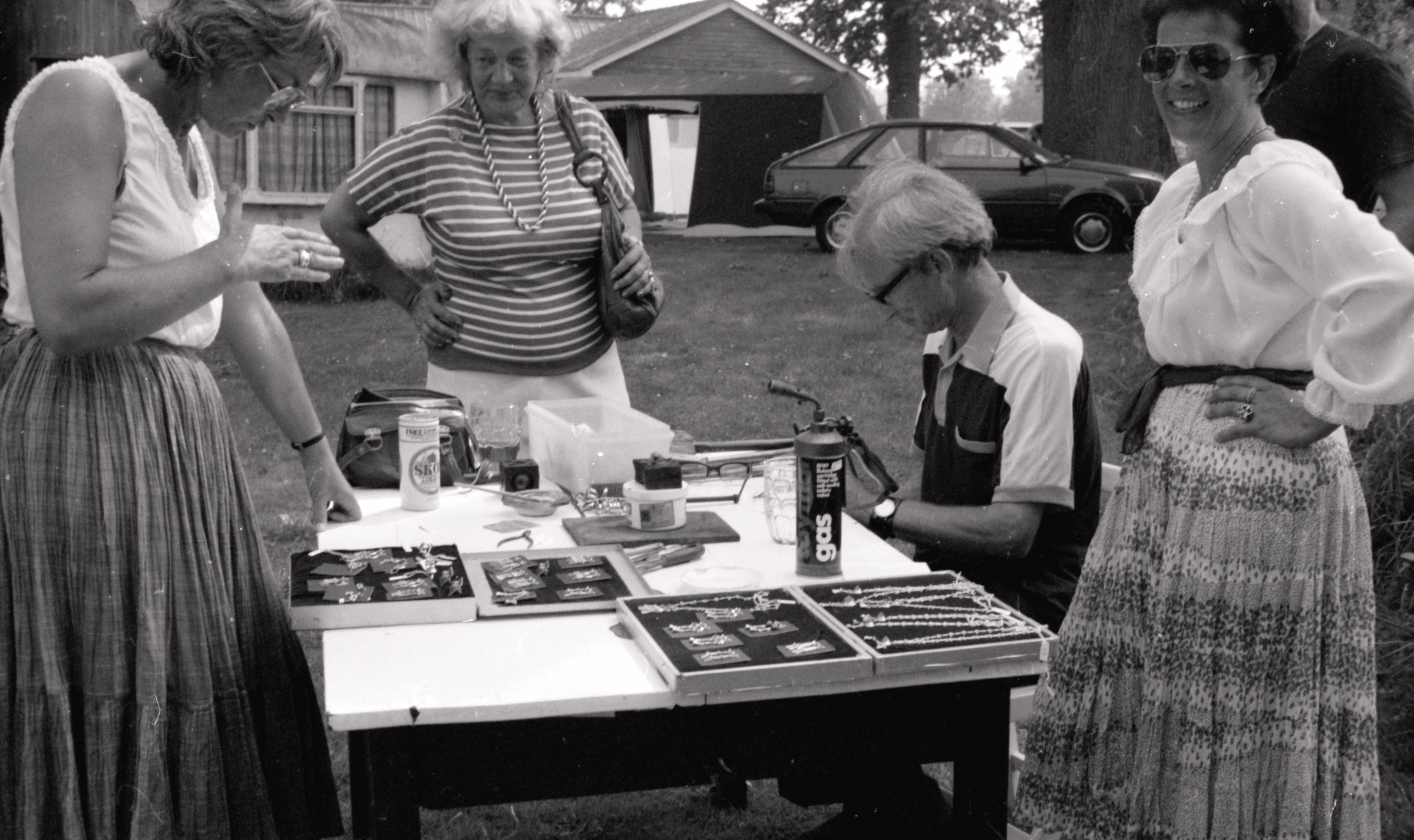 Almelijas LNPL Latviešu mājas 30. jubilejas amatnieku tirdziņš, Almelija, Herefordšīra, 1983. gada 16. vai 17. jūlijs. Sēž rotkalis Kārlis Uskalis, pa labi viņa sieva Ilona Uskalis, pa kreisi Ilonas māte Austra Korps un Vija Zauers.