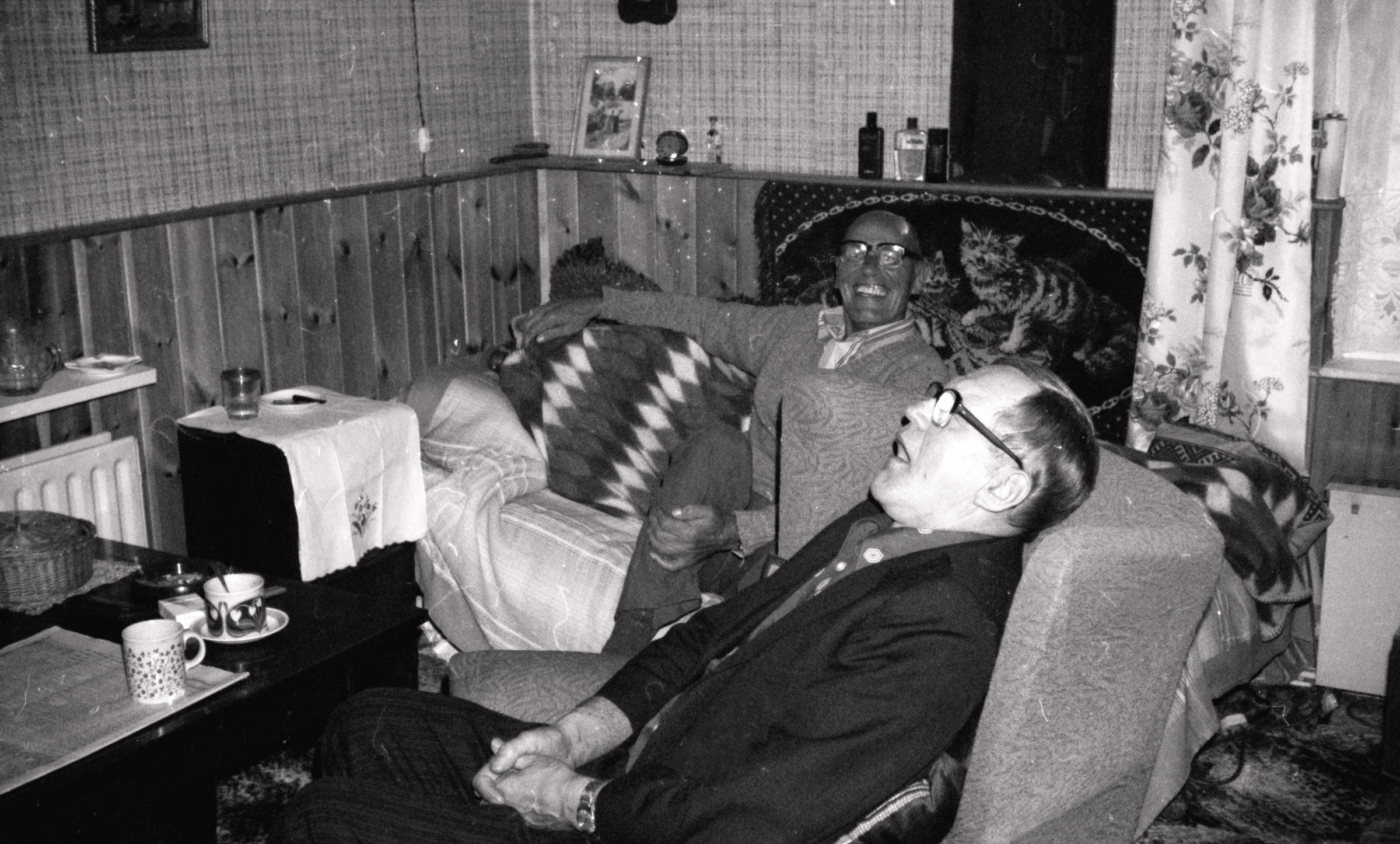 Sēžot aizsnaudies Ēvalds Petrovs, aizmugurē smaida kokļu meistars Vitolds Rūniks, LNPL Latviešu māja Almelijā, Herefordšīrā, 1984. gads.